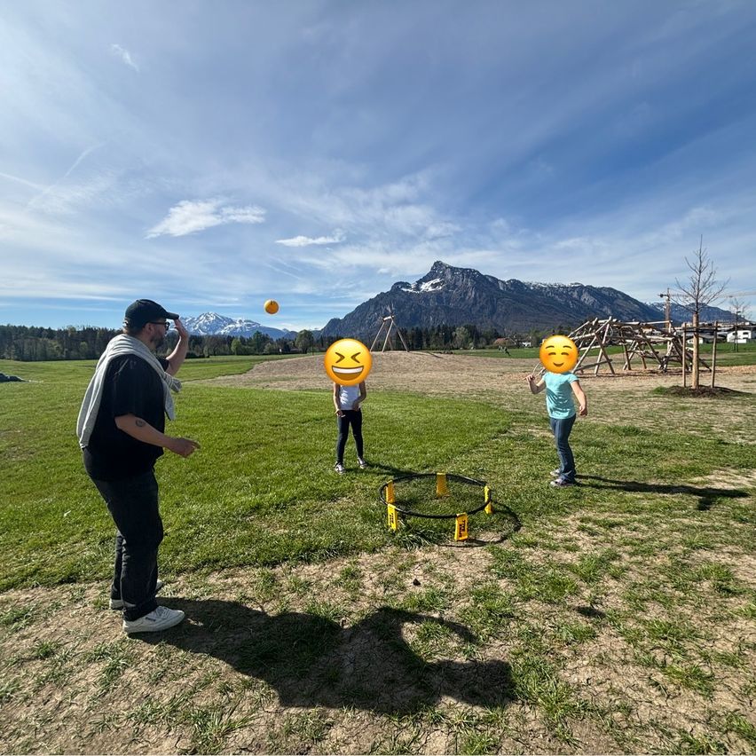 Ein Mann und zwei Kinder spielen Ball auf einem Feld. Der Mann wirft den Ball auf die Kinder, die lächeln.
