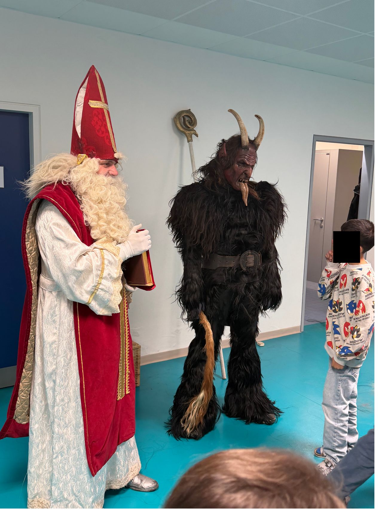 Ein Mann in einer Santa-Claus-Verkleidung steht neben einer Person in Teufelskostüm, hält ein Buch in der Hand und unterhält sich mit einem Kind in einem Raum mit blauem Boden.