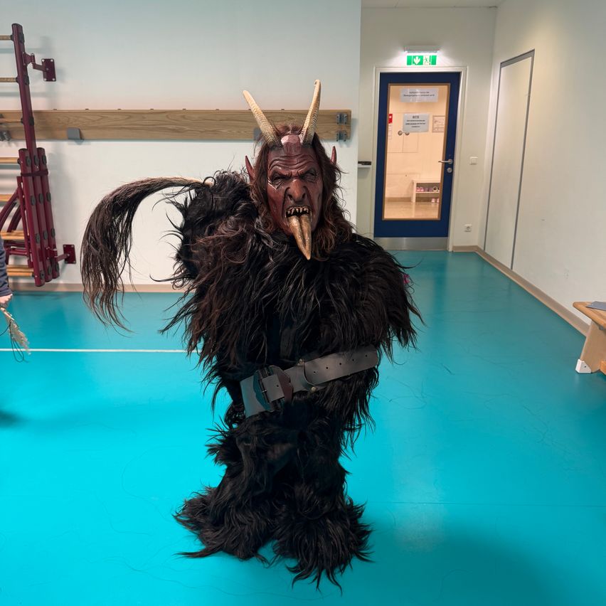 Eine Person in einem pelzigen Kostüm mit Hörnern und einem langen Rüssel steht in einer Turnhalle. Dahinter ist eine Tür mit einem Schild zu sehen, und eine Leiter befindet sich links. Der Boden ist blau.