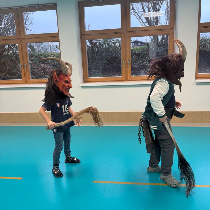 Zwei Personen in Teufelskostümen stehen in einem Raum mit blauen Boden und großen Fenstern. Einer hält einen langen Stock, der andere hat eine Besen.