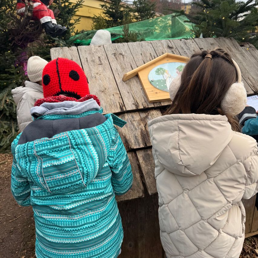 Zwei Kinder in Winterjacken betrachten eine Holzstruktur, eines trägt eine rote Marienkäfermütze. Ein Weihnachtsbaum und der Weihnachtsmann sind im Hintergrund.
