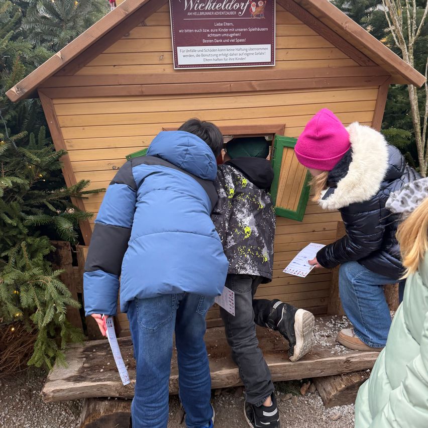 Mehrere Kinder schauen in eine Holzhütte mit einem Schild, das 'Wichteldorf' liest, und halten Papiere. Ein Kind trägt eine rosa Mütze. Sie scheinen an einer weihnachtlichen Aktivität teilzunehmen.