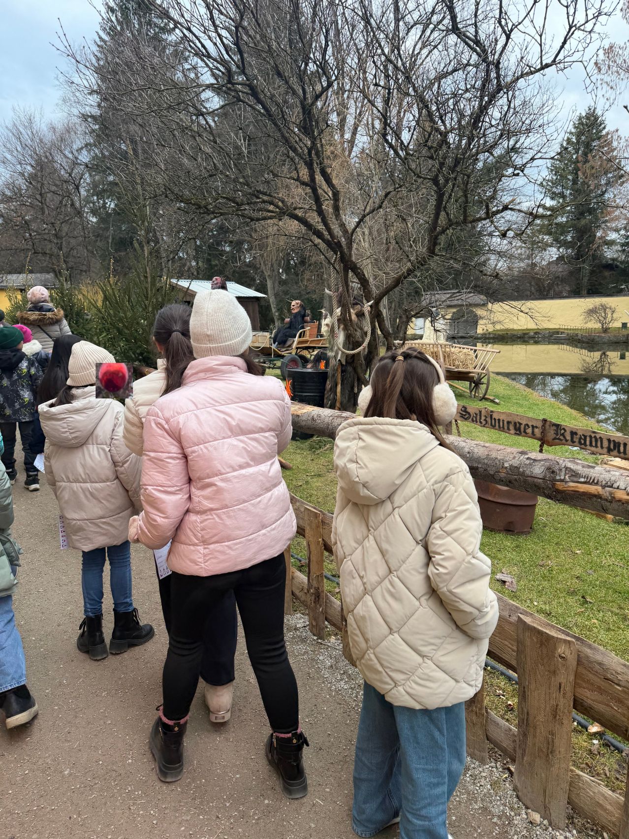 Eine Gruppe von Menschen, einschließlich Kindern, steht in der Nähe eines Zauns und schaut auf etwas vor sich. Sie tragen Winterkleidung. Eine Person sitzt auf einem Karren, an dem ein Seil befestigt ist. Ein Baum steht vor ihnen und ein Gewässer dahinter.