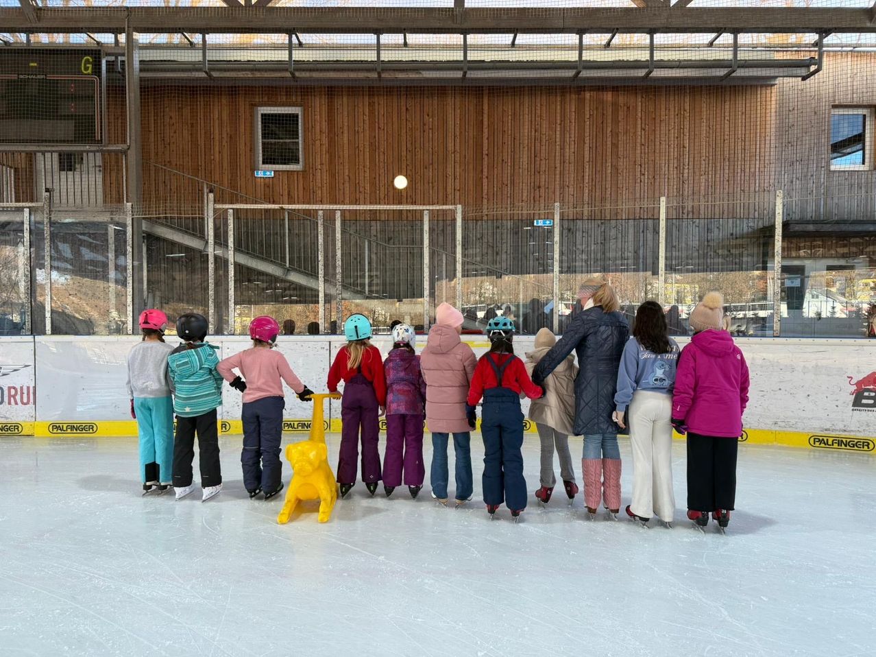 A group of children in winter clothes and helmets skate around a yellow dog on an indoor rink, with adults and a building in the background.