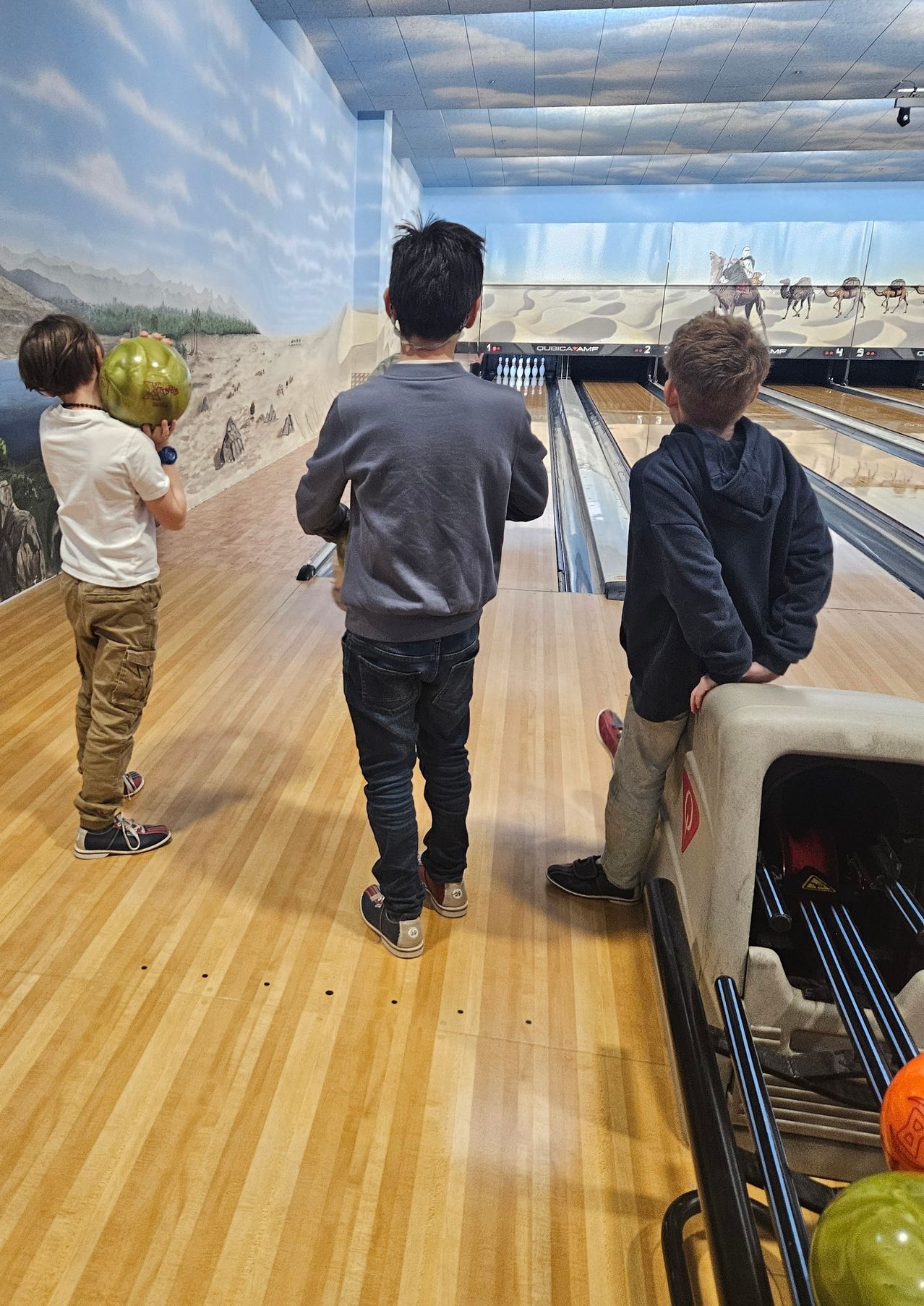 Drei Jungen sind in einer Bowlingbahn. Ein Junge hält einen grünen Ball, während die anderen beiden zusehen. Die Bowlingbahn hat Holzböden und malerische Wandgemälde.