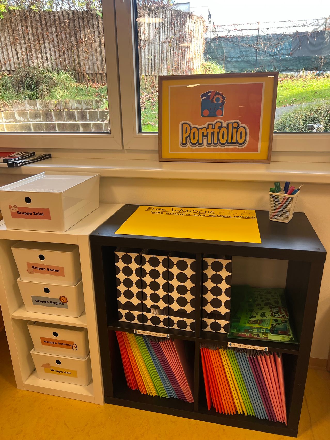 A classroom with a shelf and storage boxes labeled 'Gruppe Zeila', 'Gruppe Barbel', 'Gruppe Brigitte', 'Gruppe Sabrina', and 'Gruppe Anil'. Above it, a framed 'Portfolio' sign and a basket of pens. Outside, a grassy area with a brick wall.