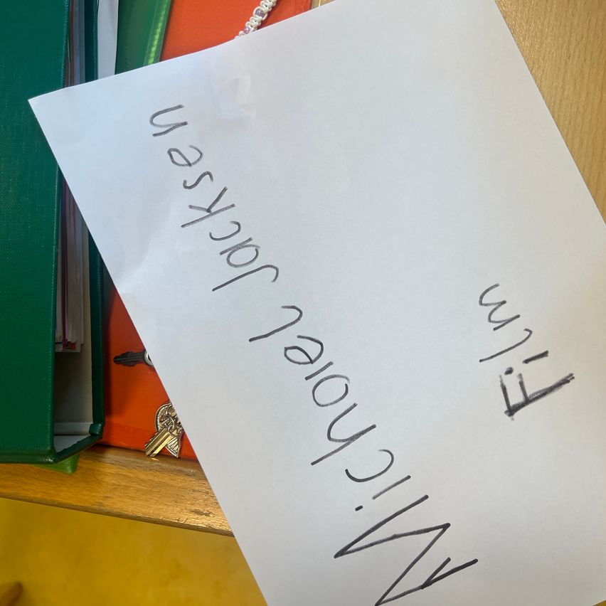 A handwritten note on white paper with the words 'November 12th' and 'Will' written in black ink. The note is placed on a wooden shelf with a green folder and keys.