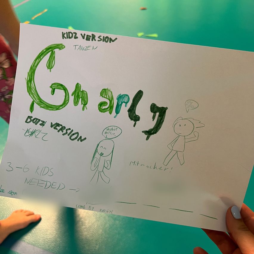 A hand holds a white sheet of paper with a drawing of two children and the text 'Gnarly' in green. The text 'KIDZ VERSION' is written in black above the drawing. The drawing includes a boy and a girl with speech bubbles. The paper also has the text '3-6 KIDS NEEDED' and 'SONG BY KATELYE'.
