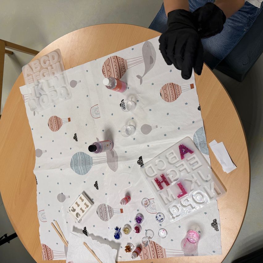 A craft table with a tablecloth featuring balloons and stars, holding various craft supplies, including a bottle, paintbrushes, and a white tray with alphabet letters. A person in black gloves is nearby.