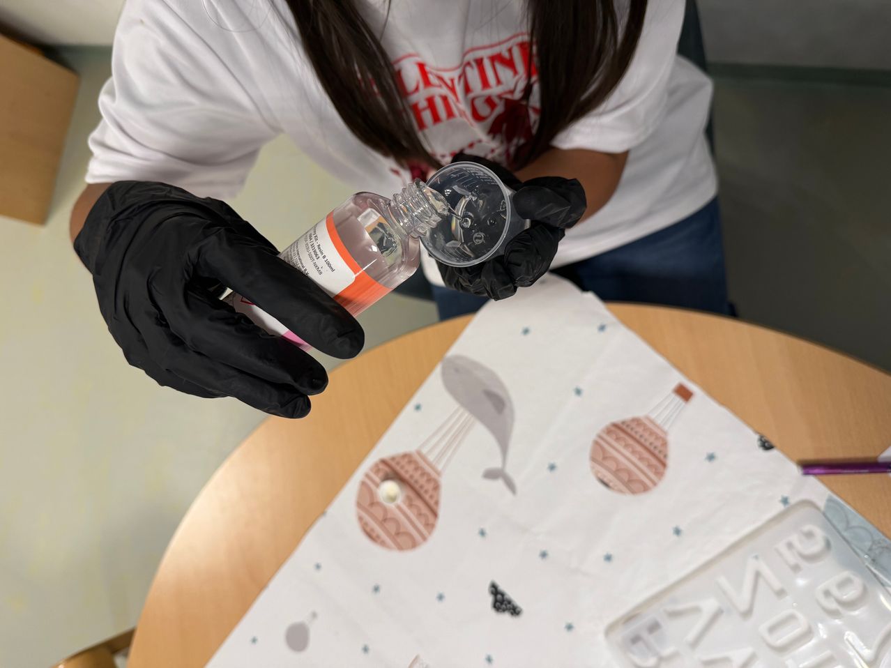 A person in a lab, wearing gloves, pours liquid from a bottle into a small container. The table has a printed cloth with balloons and a whale.