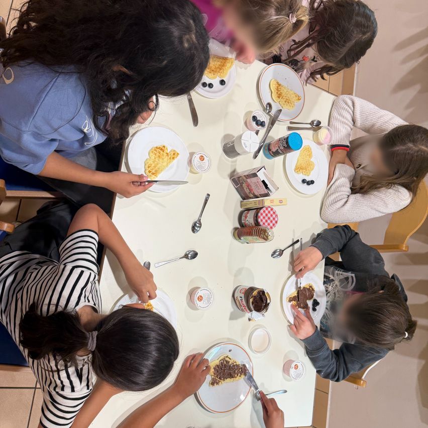 Several children sit around a table eating waffles with syrup and blueberries, some with utensils in hand. Various condiments and plates are on the table, and the children's faces are blurred.