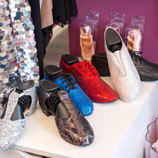 Eine Auswahl verschiedener glitzernder Schuhe in verschiedenen Farben wie Blau, Rot und Silber steht auf einem weißen Regal. Dahinter befinden sich mehrere durchsichtige Plastikbehälter mit Schuhen darin.