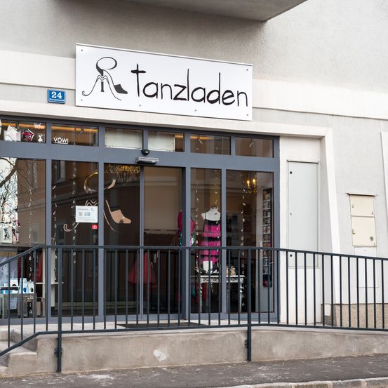 Fassade des Tanzladens mit einem Schild über dem Eingang. Das Geschäft zeigt verschiedene Artikel im Inneren, einschließlich eines Mannequins und Schmuck. Ein schwarzer Handlauf befindet sich auf den Stufen, die zum Eingang führen.