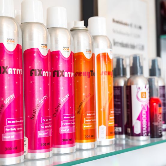 Eine Reihe verschiedener Fixative-Sprühflaschen in verschiedenen Farben, einschließlich Pink, Orange und anderen, auf einem Glasregal ausgestellt. Jede Flasche hat ein Etikett, das ihre Verwendung und ihr Volumen angibt.