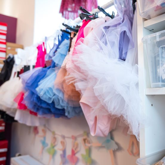 Ein Raum mit einer Wand voller bunter Balletttutus, ordentlich an Kleiderbügeln aufgehängt. An der Wand befindet sich auch ein Wandbild von Ballerinen. Im Hintergrund sind Aufbewahrungsboxen zu sehen.