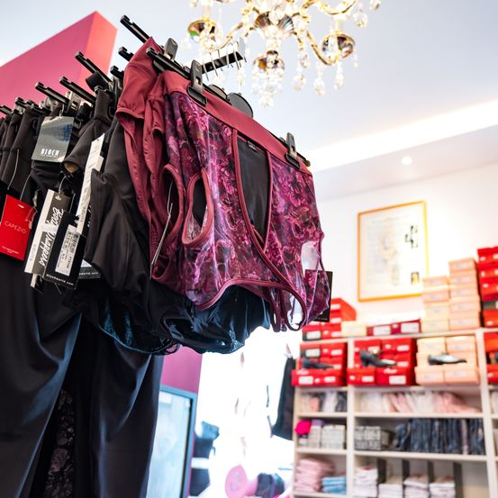 Ein Boutique zeigt verschiedene Paar Damenunterwäsche, die an Kleiderbügeln hängen. Eine Kristallleuchter hängt von der Decke. Im Hintergrund sind Regale mit ordentlich angeordneten Kartons zu sehen.