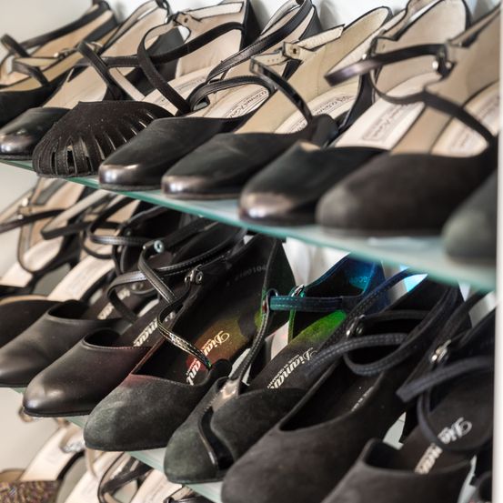 Eine Reihe von schwarzen und beigen Schuhen, hauptsächlich Absätze, auf einem Glasregal. Einige Schuhe haben Riemendesigns, während andere Schnallen haben. Die Schuhe sind ordentlich in Reihen angeordnet.