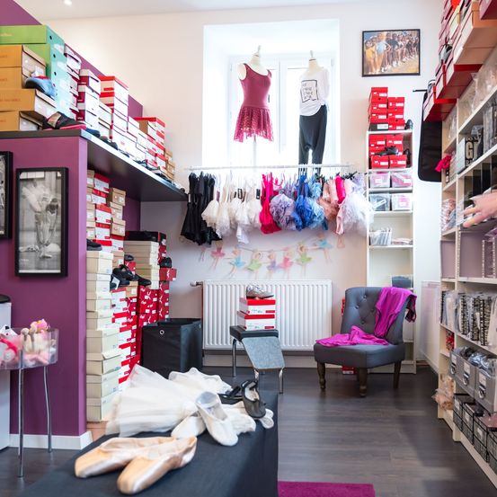 Ein Tanzstudio zeigt Regale mit verschiedenen Schuhen, Kleidern und Accessoires. In der Nähe eines Fensters befinden sich Kleider auf Mannequins. Ein Stuhl und ein Schreibtisch halten weitere Gegenstände.