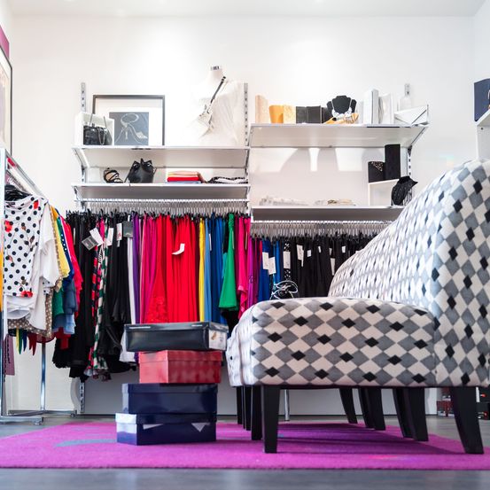 Ein Bekleidungsgeschäft mit farbenfrohen Kleidern an Kleiderbügeln, einem Regal mit verschiedenen Gegenständen und einem gemusterten Stuhl auf einem pinkfarbenen Teppich.