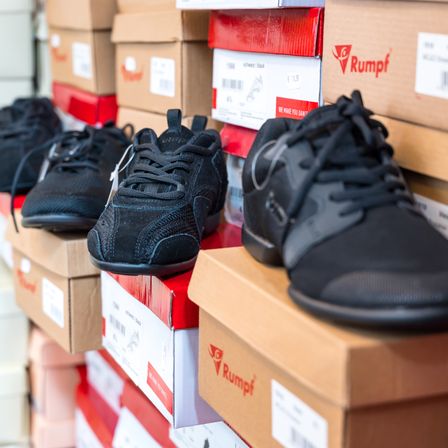 Mehrere schwarze Turnschuhe, einige in Rumpf-Kisten, sind übereinander gestapelt. Die Schuhe haben Schnürsenkel und liegen auf verschiedenen Kisten mit Barcodes und Etiketten.