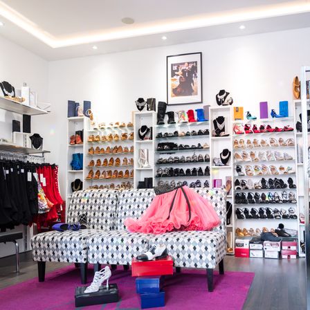 Ein Boutique-Laden mit weißen Wänden und Regalen voller Schuhe, einem Sofa mit einem rosa Tutu und Schmuck.