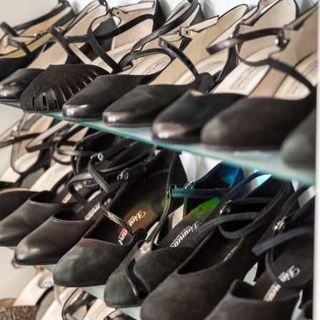 Eine Sammlung von schwarzen Schuhen, hauptsächlich aus Leder, in Reihen auf Glasregalen angeordnet. Einige haben Riemen und Schnallen, während andere flach und schlicht sind.