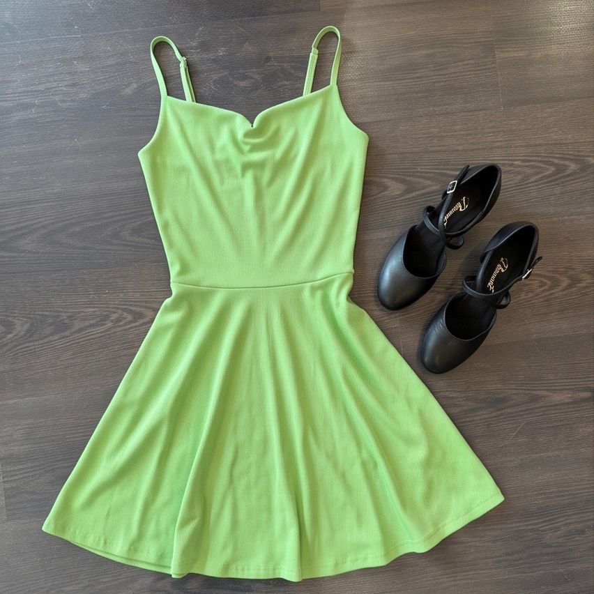 Ein lindgrünes Kleid mit V-Ausschnitt und trägerlosen Schultern liegt auf einem Holzboden neben einem Paar schwarzer Lederschuhe.