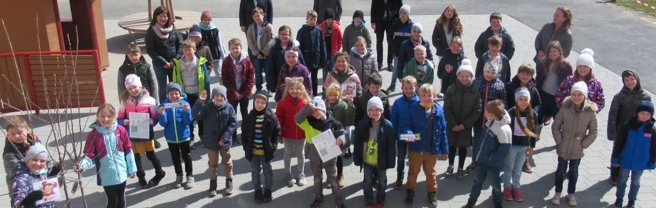 Eine Gruppe von Kindern und Erwachsenen steht in einer Reihe, hält Urkunden und lächelt für ein Foto. Die Kinder tragen Winterkleidung, einschließlich Mützen und Stiefeln. Der Hintergrund ist ein gefliester Boden.