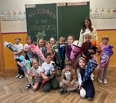 Eine Gruppe von Kindern und einer Lehrerin posiert für ein Foto im Klassenzimmer. Die Kinder halten dekorierte Kegel. Die Lehrerin steht hinter ihnen. Im Hintergrund befindet sich eine Tafel mit einer Nachricht.