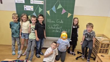 Eine Gruppe von Kindern im Klassenzimmer, einige lächeln und andere posieren für ein Foto. Ein Tafel hinter ihnen hat bunte Wimpel und die Botschaft 'Alles Gute zum Schulstart in die 2. Stufe!' darauf geschrieben.