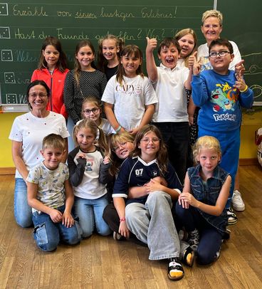Eine Gruppe von Kindern und zwei Lehrern posiert für ein Foto im Klassenzimmer. Die Kinder sitzen und stehen vor einer Tafel.