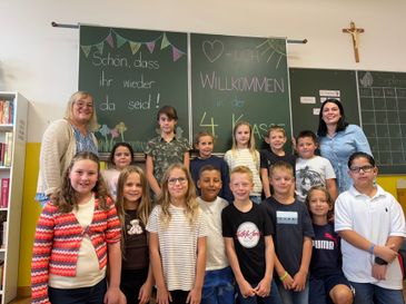 Eine Gruppe von Kindern und zwei Lehrern steht vor einer Klassentafel mit fröhlichen Dekorationen und Botschaften.