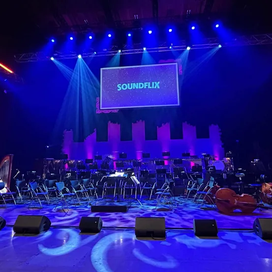 Bühne für ein Konzert vorbereitet mit Musikinstrumenten und Stühlen, große Leinwand mit dem Text Soundflix auf violettem Hintergrund, beleuchtet von blauen Lichtern.