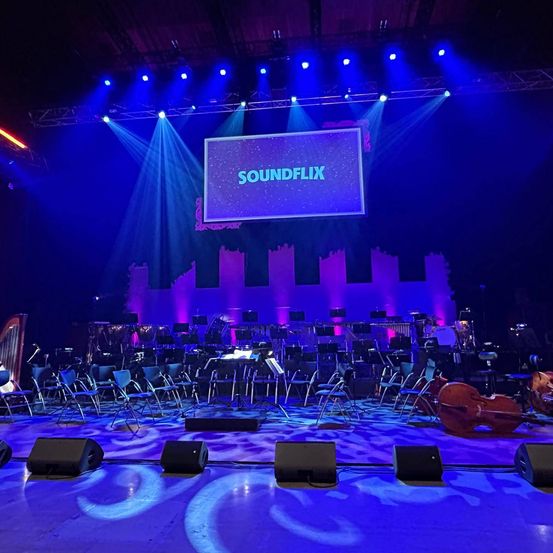 Bühne für ein Konzert vorbereitet mit Musikinstrumenten und Stühlen, große Leinwand mit dem Text Soundflix auf violettem Hintergrund, beleuchtet von blauen Lichtern.