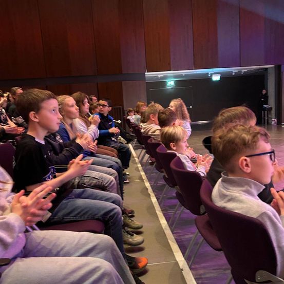Eine Gruppe von Kindern in einem Theater, die in Reihen sitzen und klatschen.