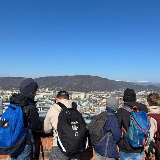 Eine Gruppe junger Schüler mit Rucksäcken, die auf einer Mauer stehen und die Stadt unten betrachten, mit einem Bergrücken und klarem blauem Himmel im Hintergrund.