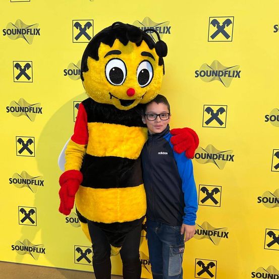 Ein Junge in einer schwarzen und blauen Jacke posiert mit einem Maskottchen, das als Biene verkleidet ist, vor einem gelben Hintergrund.