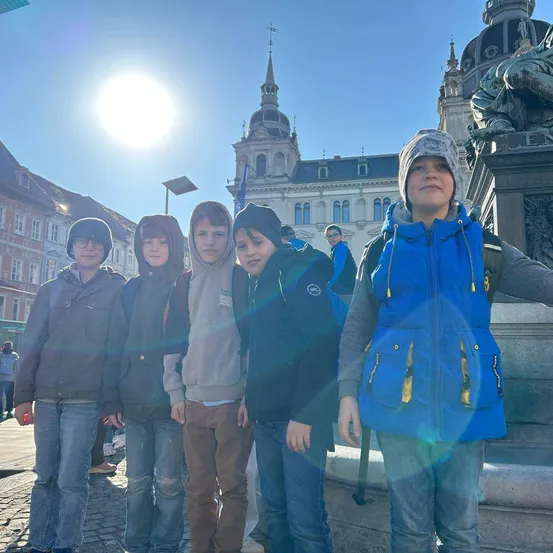 Fünf Jungen stehen vor einer Statue. Die Sonne scheint hell. Sie tragen Jacken und Jeans. Sie lächeln. Hinter ihnen steht ein Gebäude mit vielen Fenstern. In der Ferne gehen Leute spazieren.