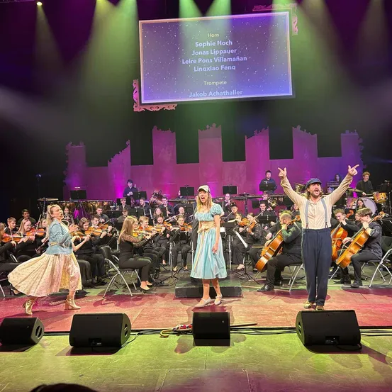 Eine Gruppe von Musikern auf der Bühne, die verschiedene Instrumente spielen. Eine Frau spielt Geige, eine andere Frau singt und ein Mann spielt Trompete. Dahinter spielt ein Orchester.