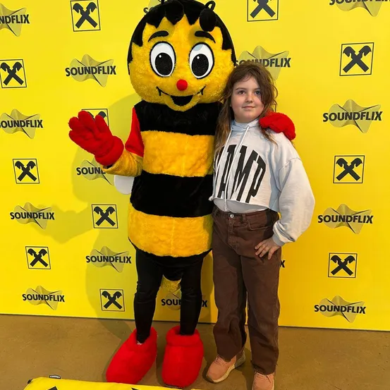 Ein junges Mädchen in einer grauen Kapuzenjacke posiert mit einem Maskottchen, einer gelben und schwarzen Biene mit roter Nase. Der Hintergrund ist eine gelbe Wand mit Logos und dem Wort Soundflix.