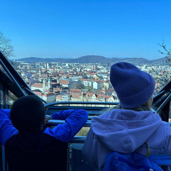 Zwei Kinder schauen aus dem Fenster einer Seilbahn auf eine Stadt mit vielen Gebäuden und Bergen in der Ferne.