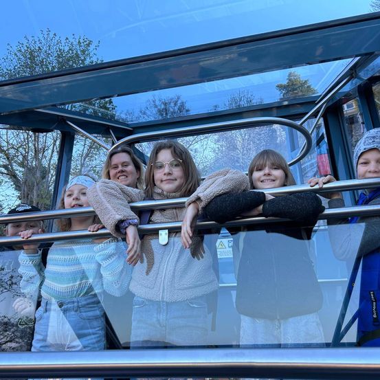 Eine Gruppe von Kindern und Erwachsenen, darunter eine Frau mit Brille, posiert in einer transparenten Kabine mit einem Metallgeländer.