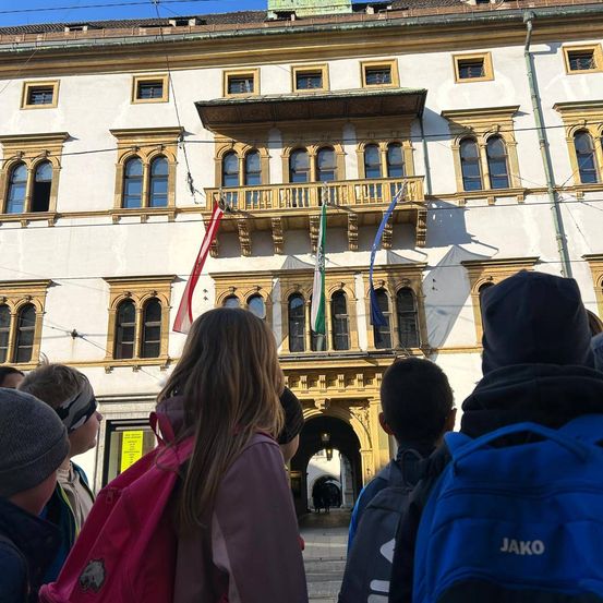 Eine Gruppe von Kindern mit Rucksäcken steht vor einem Gebäude mit Bogenfenstern und einer Balkon mit Flaggen.