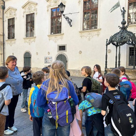 Eine Gruppe von Schülern mit Rucksäcken steht vor einem weißen Gebäude mit verzierten Fenstern und einer Lampe, die an der Wand hängt.