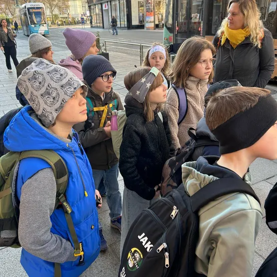 Gruppe von Schülern, die einem Lehrer auf der Straße zuhören. Sie tragen alle Winterkleidung und haben Rucksäcke dabei.