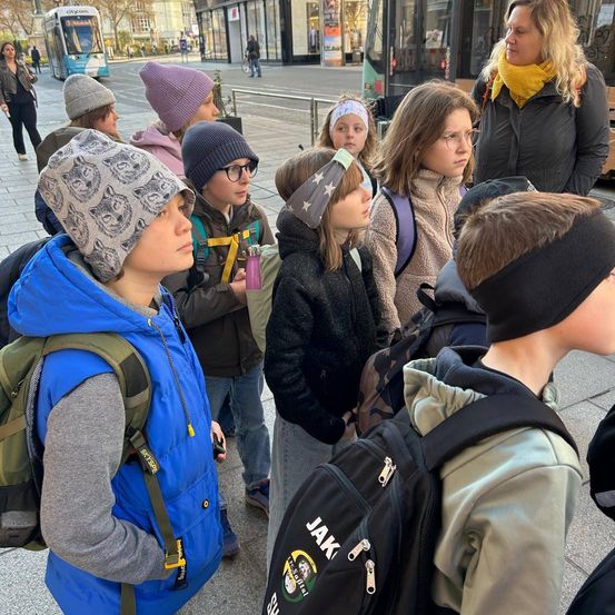 Gruppe von Schülern, die einem Lehrer auf der Straße zuhören. Sie tragen alle Winterkleidung und haben Rucksäcke dabei.