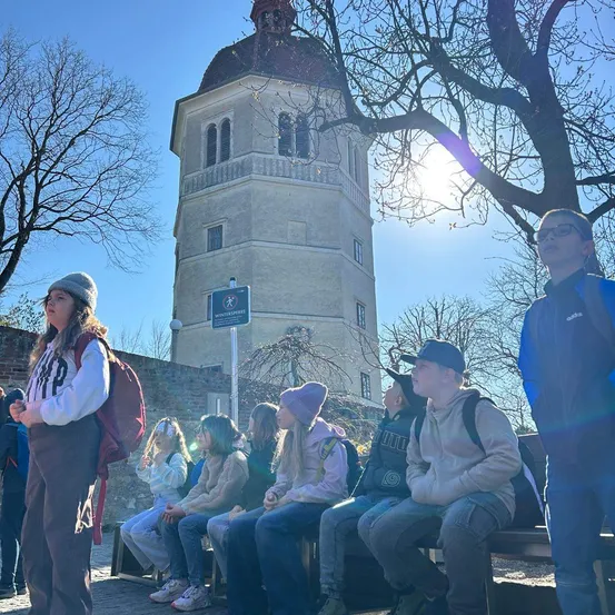 Eine Gruppe von Menschen, wahrscheinlich Schüler, sitzt auf einer Bank vor einem historischen Gebäude, wobei eine Person in der Nähe eines Baumes steht.