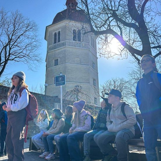 Eine Gruppe von Menschen, wahrscheinlich Schüler, sitzt auf einer Bank vor einem historischen Gebäude, wobei eine Person in der Nähe eines Baumes steht.
