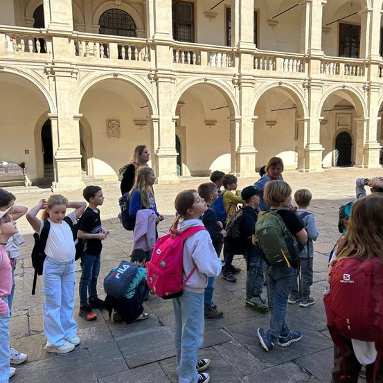 Eine Gruppe von Schulkindern mit Rucksäcken versammelt sich vor einem prunkvollen Gebäude mit Bögen und Säulen. Einige schauen nach oben, während andere miteinander sprechen.
