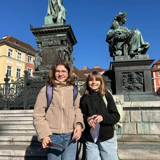 Zwei Mädchen in Jacken stehen vor einer Statue in einer Stadt. Sie lächeln und halten Papiere in der Hand. Hinter ihnen steht eine große Statue mit einem Schild, das 'Mur' besagt. Im Hintergrund sind Gebäude und ein Zaun zu sehen.